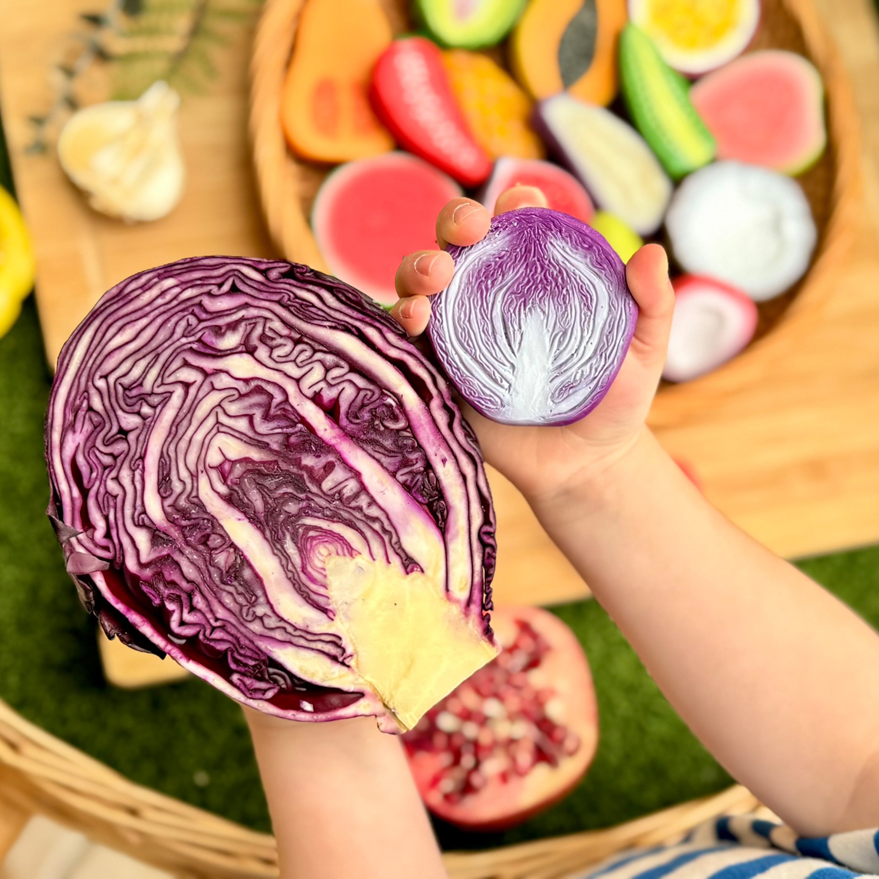 Child holding a purple cabbage with a wooden board of colorful play food in the background.