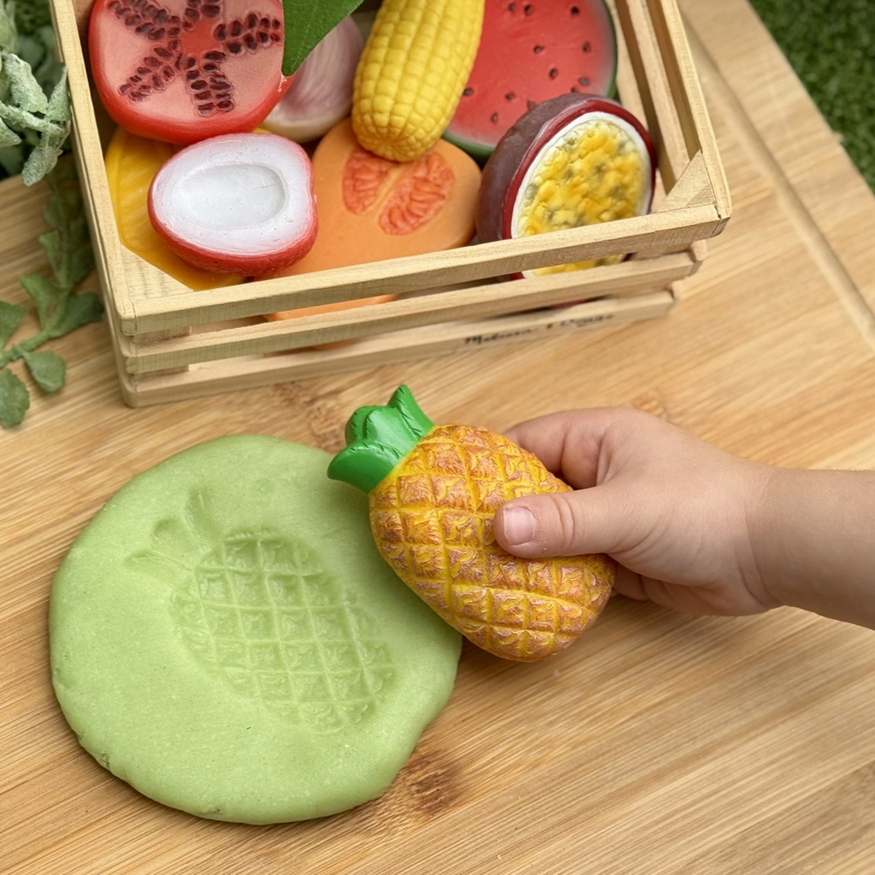 Playdough with pineapple shape held by a hand, next to a wooden crate filled with toy fruits on a wooden surface.