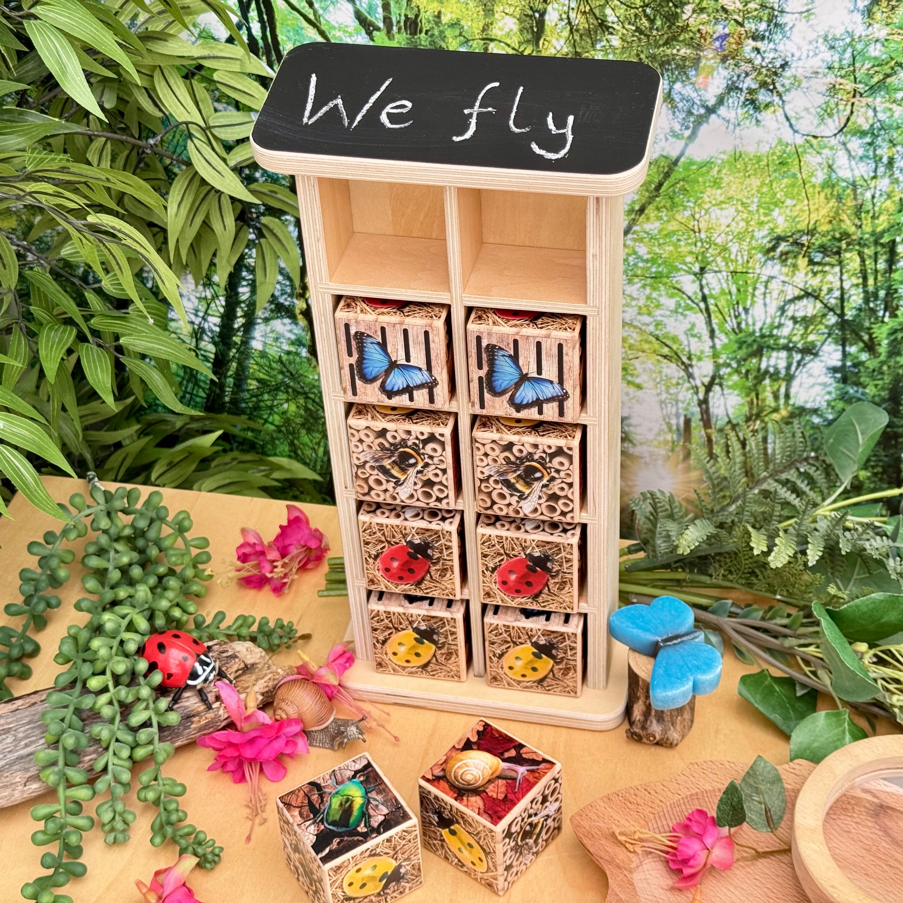 Wooden insect habitat with small boxes labeled with insects, surrounded by greenery.